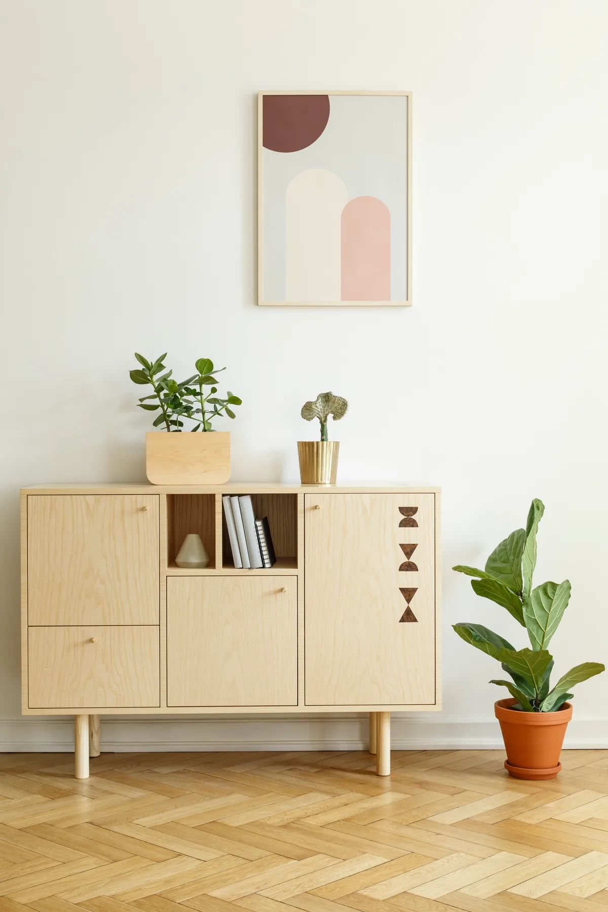 Die klaren Linien des nordischen Designs lassen sich mit wenigen, ausgewählten Dekoelementen auflockern. Vor einer weissen Wand mit einem Bild steht eine Kommode aus Holz neben einer Pflanze in einem Wohnzimmer.